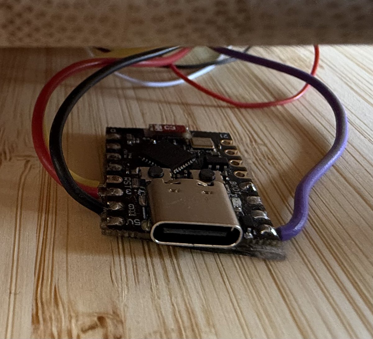 Macro of a small black ESP32-C3 Super Mini board with a USB-C port and RST, BOOT and 5V pin labels, wired to a harness of coloured jumper wires