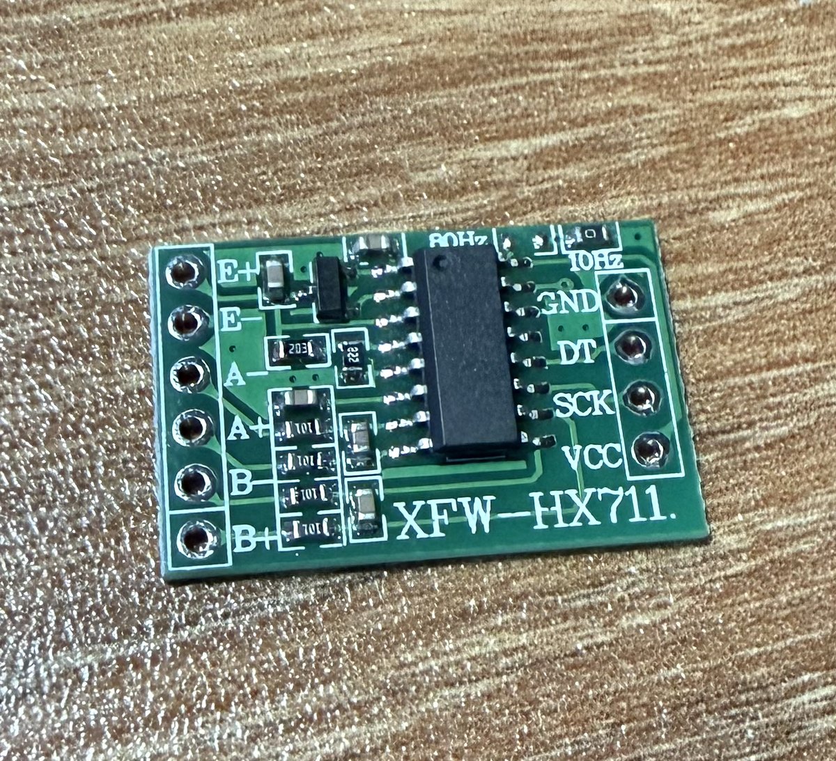Macro of a green HX711 breakout board labelled XFW-HX711, with bridge inputs E+, E−, A−, A+, B−, B+ on one side and GND, DT, SCK, VCC plus 10Hz/80Hz rate jumpers on the other